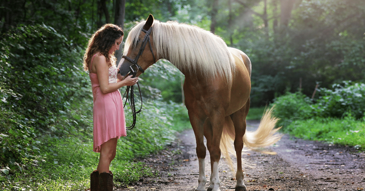 Pet-friendly rehab: Woman in pink dress gently holding a horse with a blonde mane on a wooded path. Animal therapy concept.