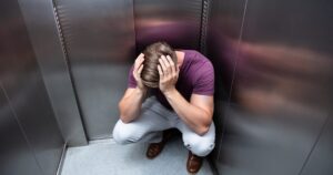 Man crouched in a small elevator with his head in his hands, looking distressed.