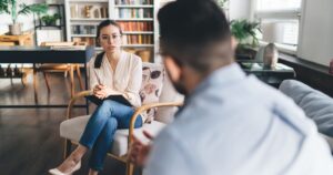 Therapist wearing glasses sits on a chair with a notebook, speaking to a client in a cozy, book-filled living room.