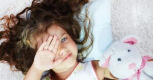 - Tennessee Behavioral Health Young girl with curly hair lying on a soft rug, hand partly covering her face, beside a white plush bunny with pink ears and ribbon.