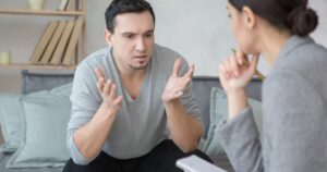 Man on a sofa speaks with animated hand movements, facing a woman who is listening and taking notes in a living room setting