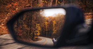 Winding autumn road seen through a car's side-view mirror, surrounded by orange and yellow trees.