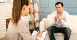 Therapist taking notes on a clipboard while speaking with a man seated on a sofa in a therapy room.