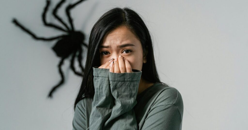 Woman in a green top covers her mouth with sleeves, looking anxious; a large spider shadow on the wall behind her.