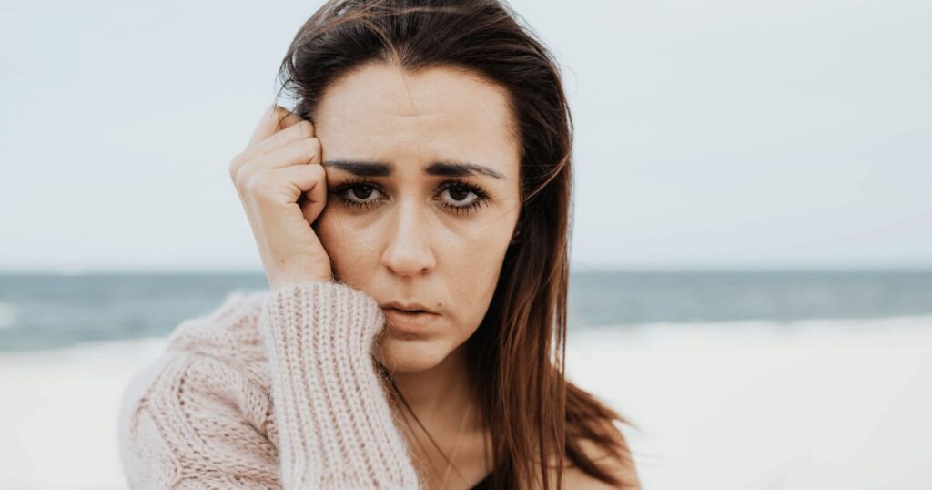 Close-up of a woman at the beach with a worried expression, hand pressed to her cheek in a pale sweater.