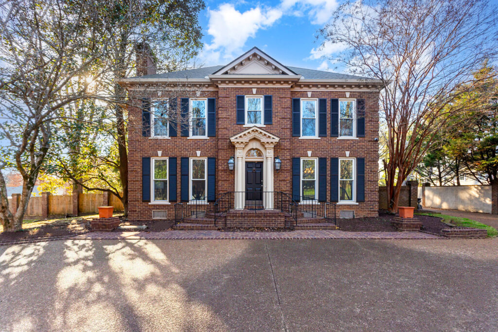 005_ndp-120623-e-00-edit - Tennessee Behavioral Health Front view of a brick two-story house with black shutters, white trim, and a decorative arched entryway accessed by small steps with an iron railing. Trees frame the scene on a sunny day.