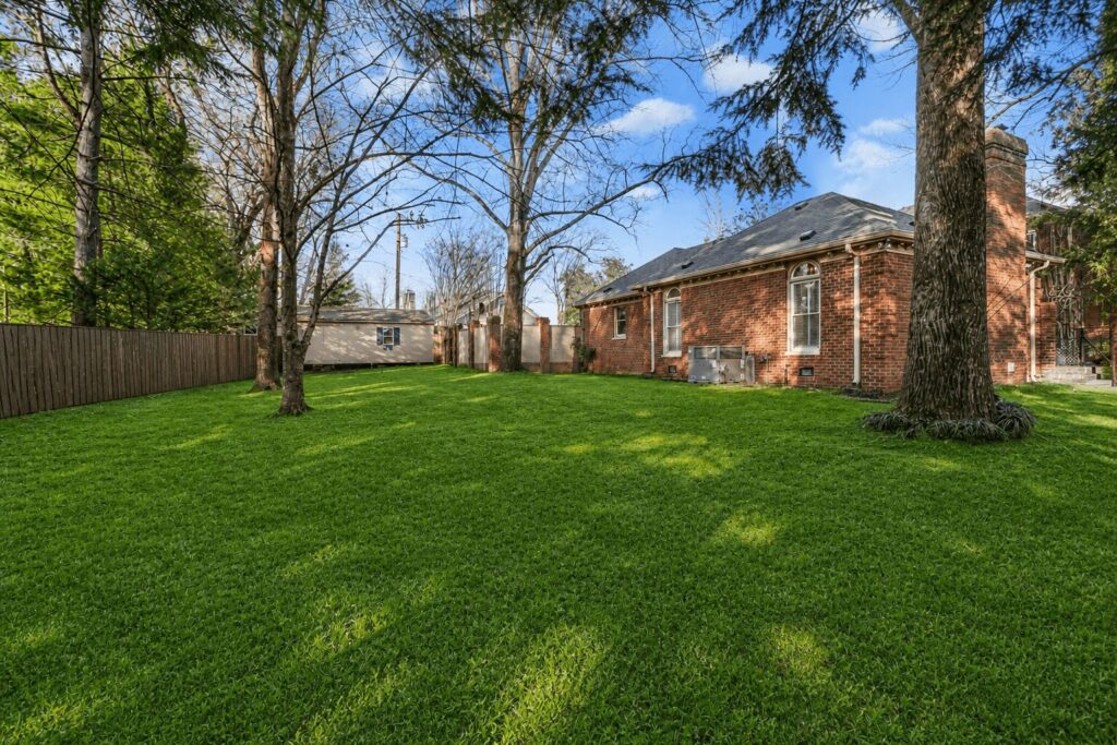 8395f397-ca64-471b-aeb6-737c56d1fac4 - Tennessee Behavioral Health Brick house facing a lush green backyard with tall trees, a wooden fence on the left, and a small shed in the distance
