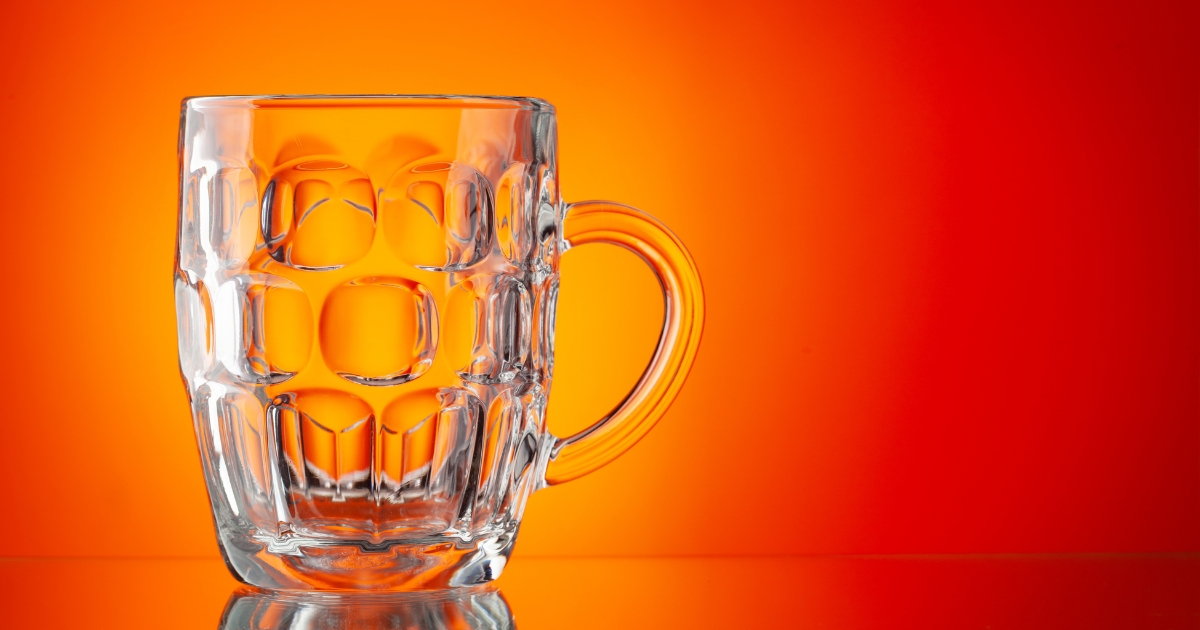 Clear glass beer mug with a curved handle set against an orange gradient background, reflecting light on the glass surface.
