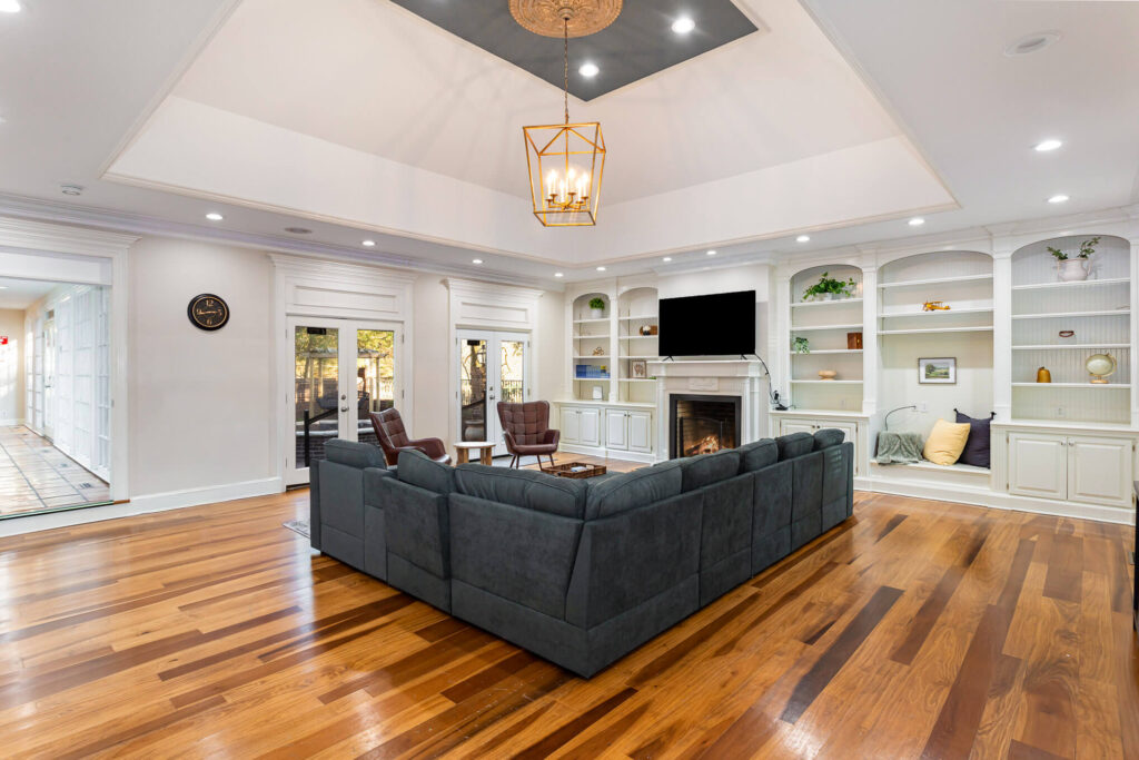 tbh-addiction-treatment-center-living-room1 - Tennessee Behavioral Health Spacious living room with a dark gray sectional sofa around a wooden coffee table, white built‑in shelves, a fireplace with a mounted TV above, and a gold geometric chandelier overhead.