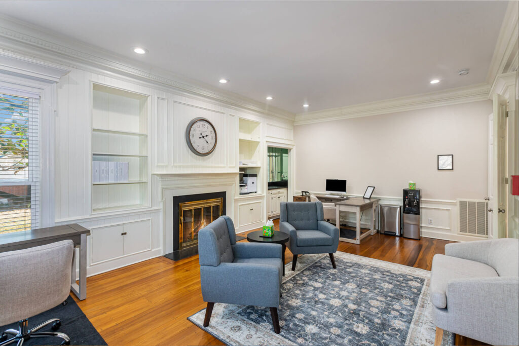 tbh-addiction-treatment-center-living-room8 - Tennessee Behavioral Health Bright home-office lounge featuring white paneled walls, built-in shelves, a fireplace, and a wall clock, with blue armchairs and a rug.