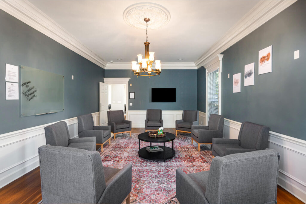 tbh-addiction-treatment-center-meeting-room2 - Tennessee Behavioral Health Conference room with blue walls and white trim, gray chairs arranged in a circle around a black coffee table under a chandelier.