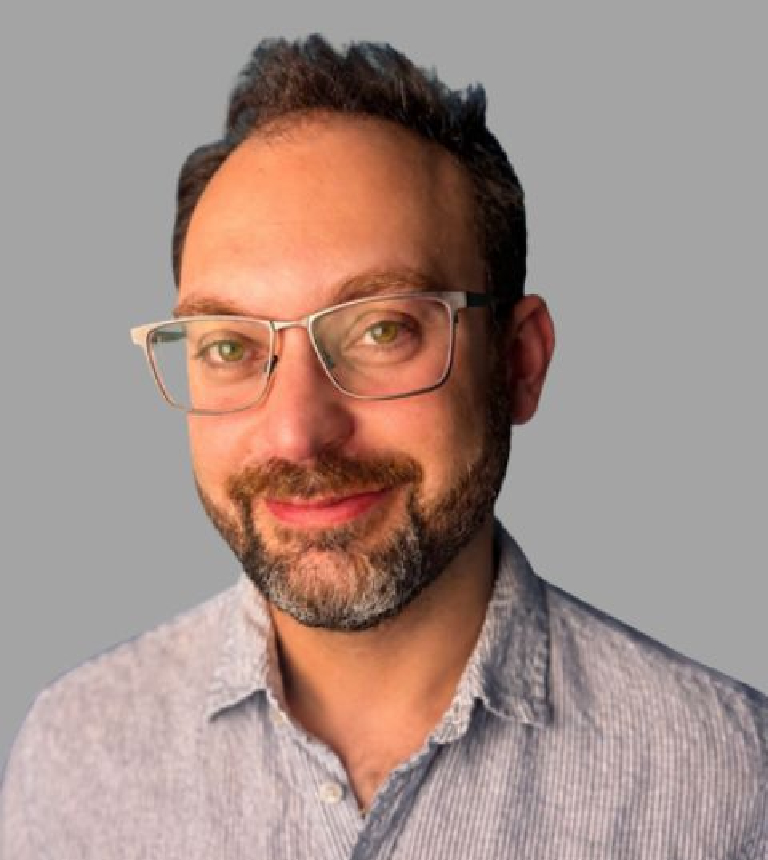 Portrait of a smiling man with glasses and a beard, wearing a light blue striped shirt, against a gray background.
